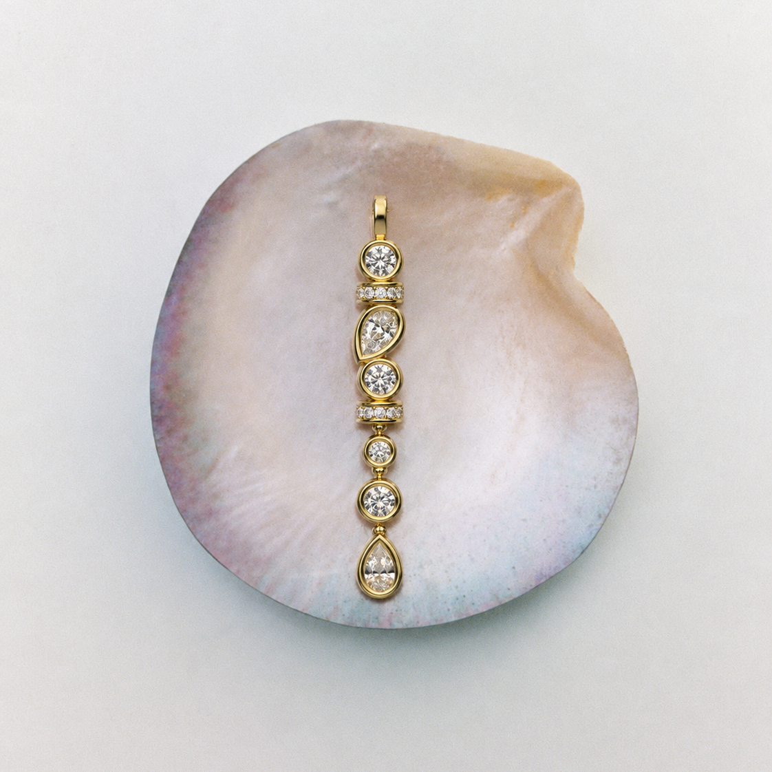 A gold pendant with five clear round-cut gemstones set in a vertical line, displayed on a large iridescent mother-of-pearl clam shell against a light gray background.