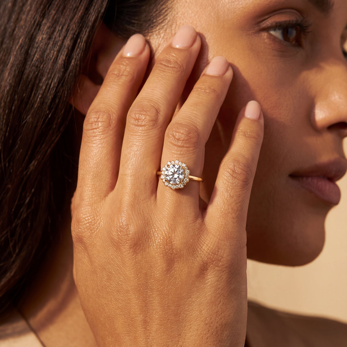 A close-up lifestyle shot of a woman wearing a yellow gold halo engagement ring with a round-cut diamond. The ring is shown on her finger, with her hand resting against her cheek, set against a warm beige background.