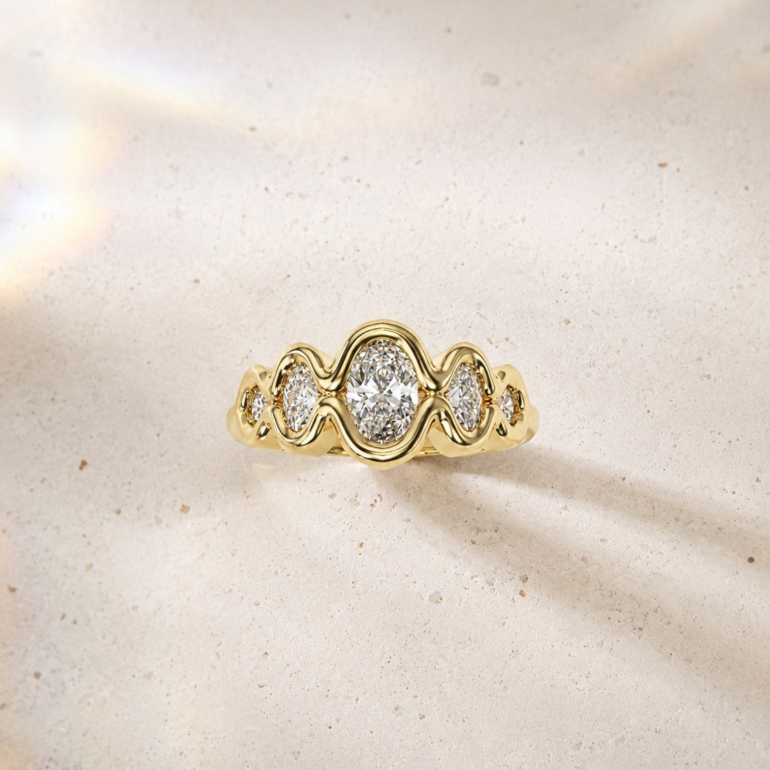 Yellow gold Flow diamond engagement ring with oval and round diamonds on a neutral beige background.