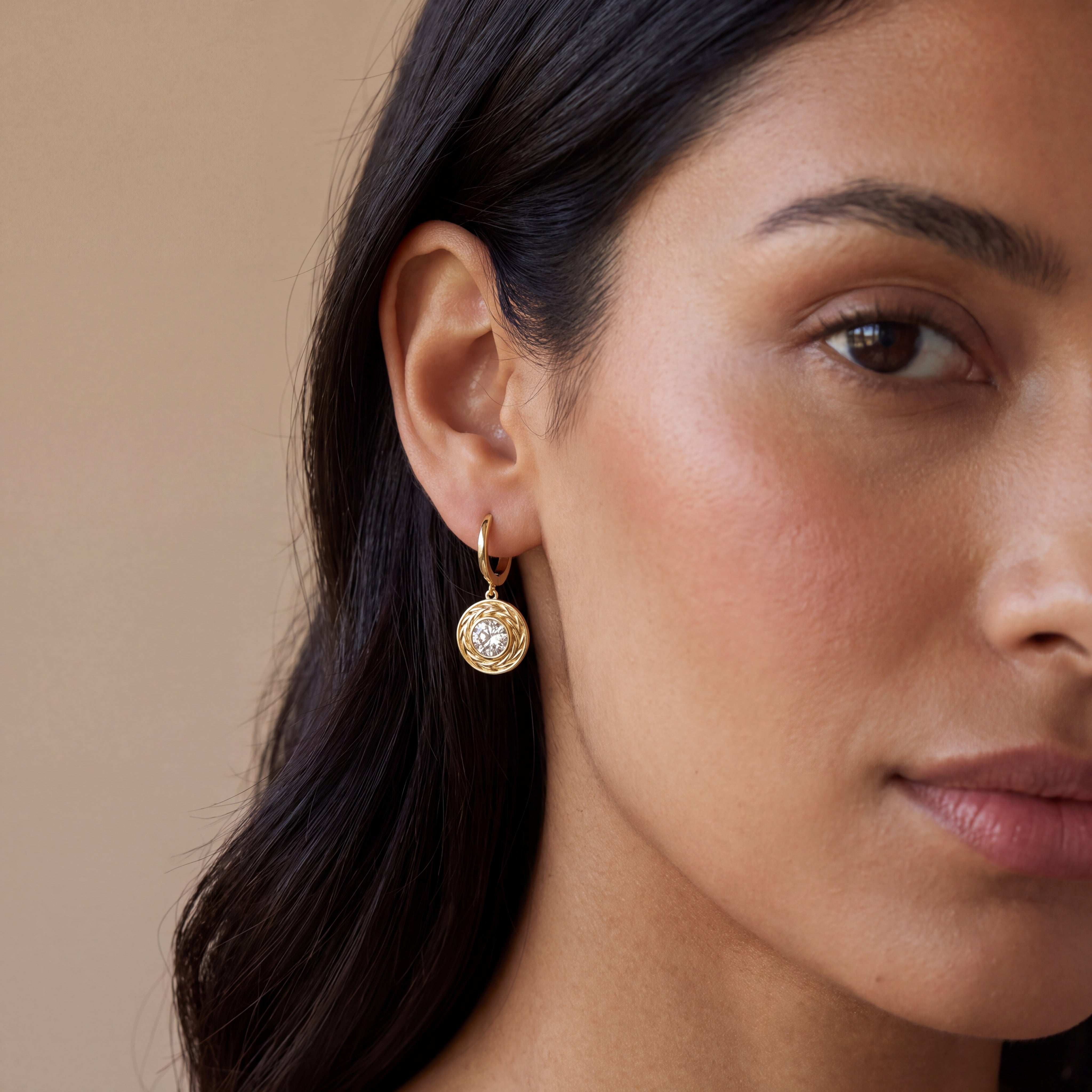 A close-up of a woman's ear wearing a small gold hoop earring with a round bezel-set clear gemstone, against a warm beige background.