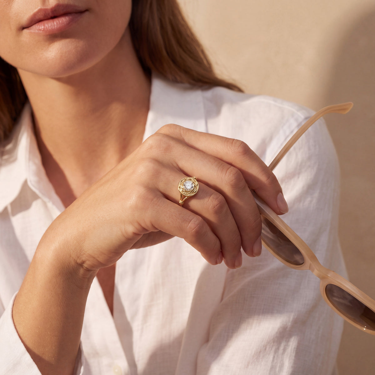 A close-up lifestyle shot of a woman wearing a gold ring with a clear round gemstone on her finger, holding a pair of light wood-framed sunglasses. She is wearing a white linen shirt against a warm beige background.