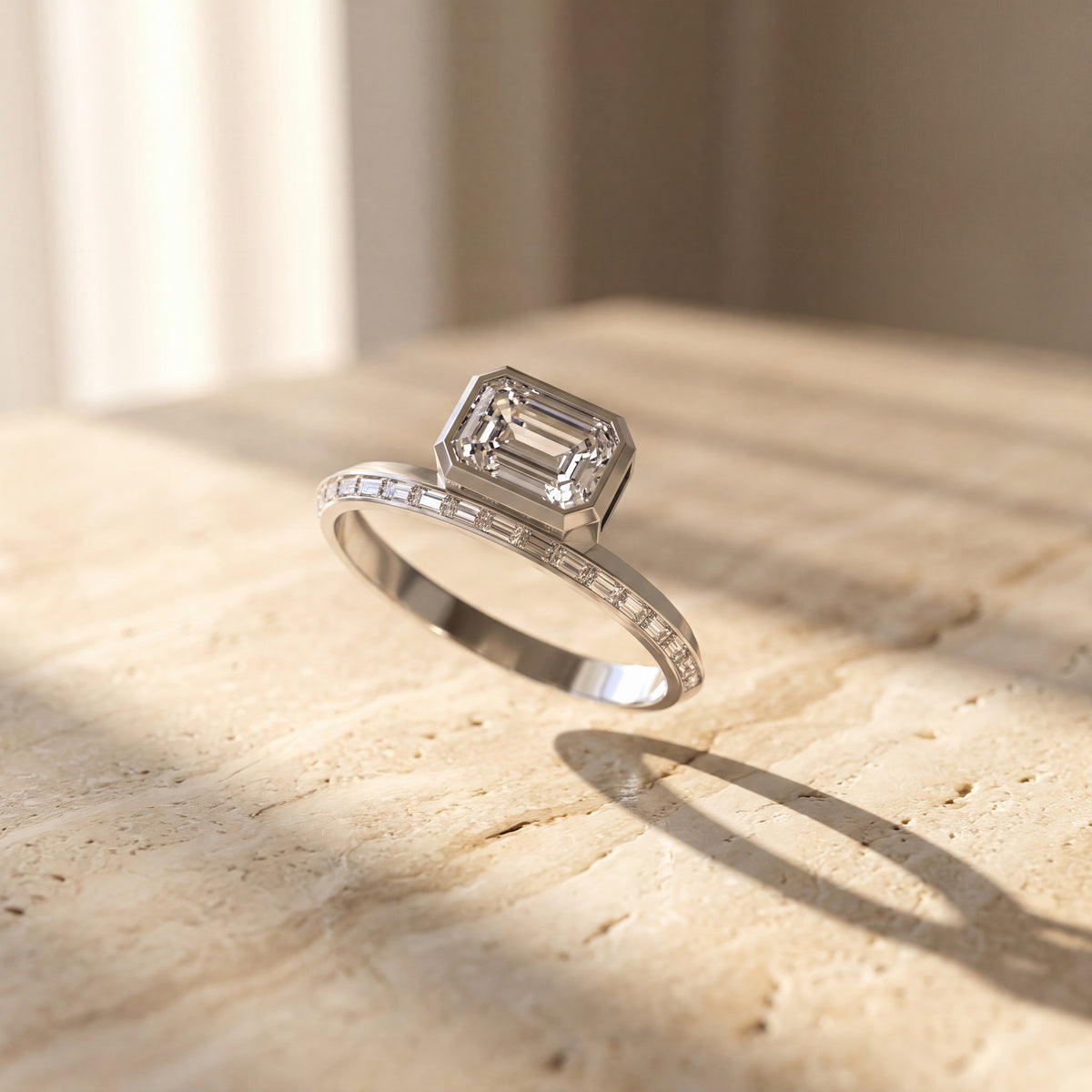 Emerald-cut diamond engagement ring with a baguette diamond knife-edge band in white gold, photographed on a textured beige stone surface.
