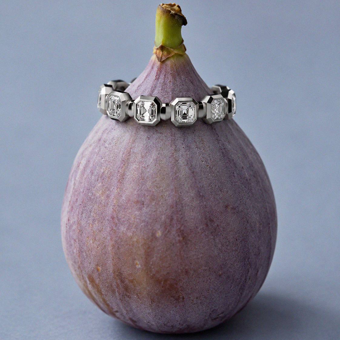 White gold Asscher-cut diamond eternity ring displayed around a fresh purple fig on a soft blue background.
